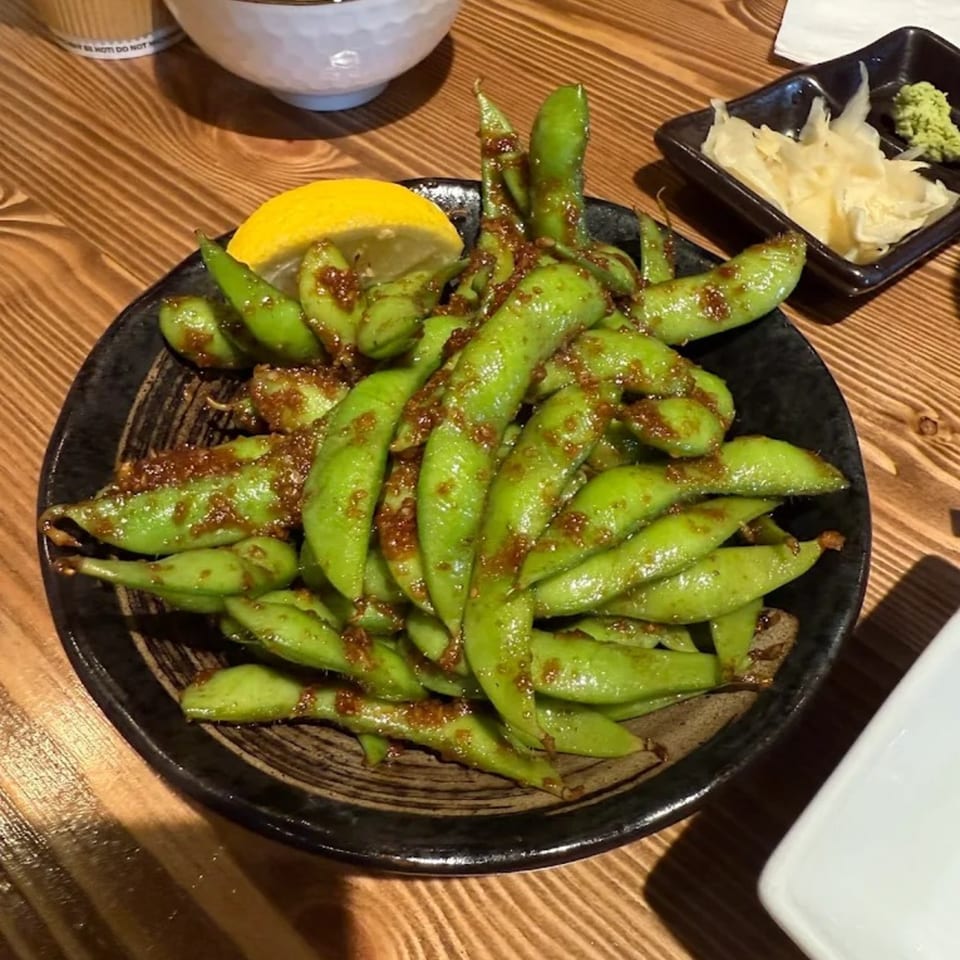 A black bowl of edamame with sauce and a lemon wedge on a wooden table.