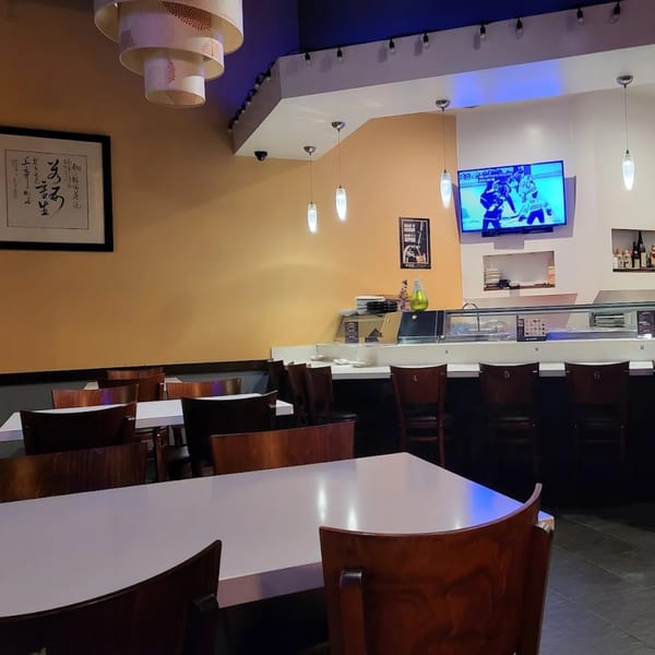Interior of Niko Niko Sushi with dining tables, a sushi bar, and a TV showing a hockey game.
